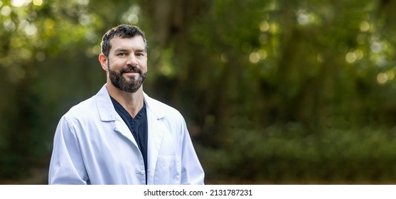 A Doctor With Dark Hair And A Beard In Black Scrubs And A White Lab Coat Standing Outside With Copy Space.