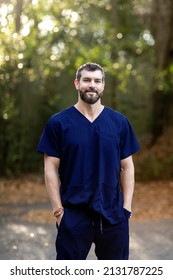 A Doctor With Dark Hair And A Beard In Navy Blue Scrubs Standing Outside In A Natural Green Environment.