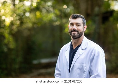 A Doctor With Dark Hair And A Beard In A White Lab Coat Standing Outside In A Natural Green Environment