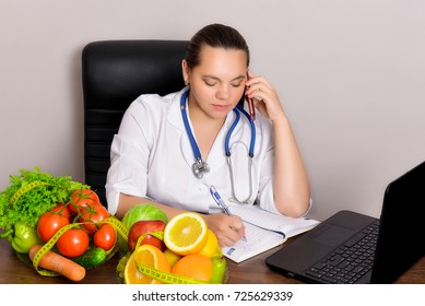 The Doctor Dietician Prescribes A Prescription For Weight Loss In The Notebook, Talking To The Patient Over The Phone. A Doctor At The Office In The Work Process On The Table Has A Lot Of Healthy Food