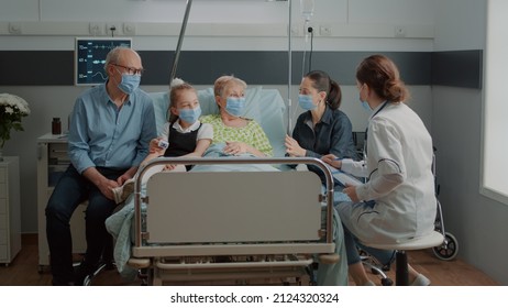 Doctor Discussing About Treatment With Ill Patient At Visit With Family During Coronavirus Pandemic. Physician Giving Medical Advice To Old Woman And Relatives With Face Mask In Hospital Ward.