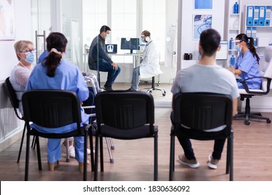 Doctor Discussing With Patient In Hospital Examination Room Wearing Face Mask Against Coronavirus And Group Of People In Clinic Lobby. Social Distancing, Medic, Covid-19, Man, Worried,