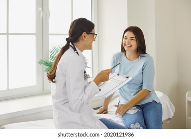 Doctor discussing treatment with cheerful smiley female patient. Happy physician and young woman talking and laughing sitting on examination bed in modern clinic or hospital - Shutterstock ID 2020121936
