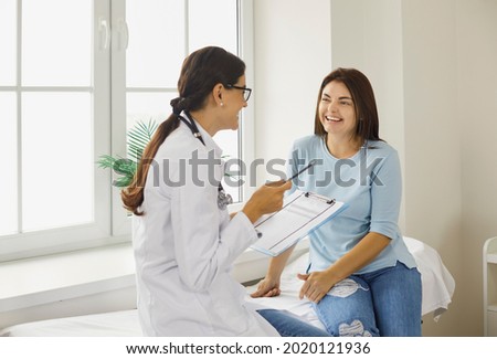 Doctor discussing treatment with cheerful smiley female patient. Happy physician and young woman talking and laughing sitting on examination bed in modern clinic or hospital