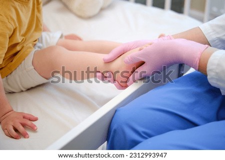 Doctor does gymnastics and foot massage to a toddler baby. Nurse in uniform doing orthopedic leg exercises to the child. Kid aged about two years (one year eleven months)