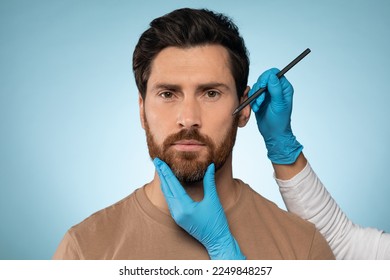 Doctor drawing marks male face for cosmetic surgery operation over blue studio background  Handsome guy getting ready for aestetic operation face lifting