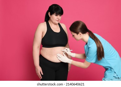 Doctor Drawing Marks On Obese Woman's Body Against Pink Background. Weight Loss Surgery