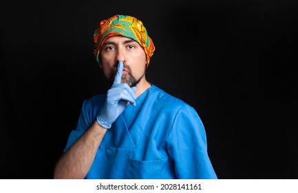 A Doctor Dressed In A Blue Hospital Uniform, Wearing An Ethnic Surgical Cap, Dark And Bearded, Asks For Silence With His Index Finger Over His Lips. A Black Background And An Area Of Copy Space 