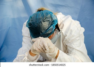 Doctor During The Coronavirus Pandemic. The Doctor Fell Asleep At The Workplace From Fatigue With Patients With Coronavirus. A Tired Doctor In A Protective Suit Prays With His Head In His Hands.