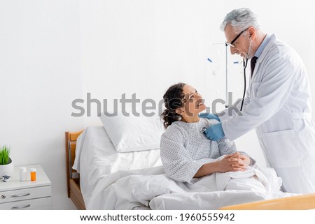 doctor examining cheerful african american woman with stethoscope in hospital
