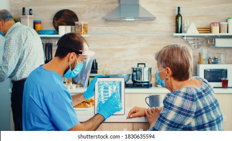 Doctor Examining Patient X-ray On Tablet Pc During Home Visit Wearing Face Mask. Elderly Person Medical Radiography By Male Nurse Discussing Disease Prevention, Examining Retired Senior Woman.