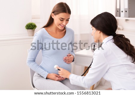 Doctor Examining Pregnant Woman Holding Stethoscope Near Belly Listening Baby's Heartbeat In Office. Pregnancy Checkup
