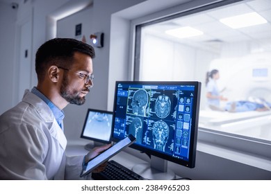 Doctor examining X-ray images on display in MRI control room while in background nurse preparing the patient for examination test.