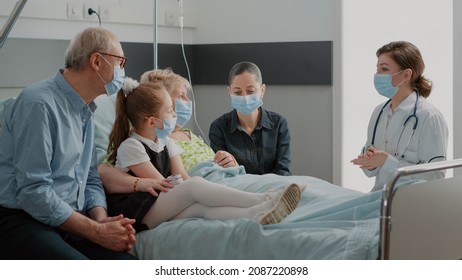 Doctor Explaining Diagnosis To People In Hospital Ward During Covid 19 Pandemic. Physician Talking About Disease With Patient And Family In Visit, Wearing Face Mask At Consultation.