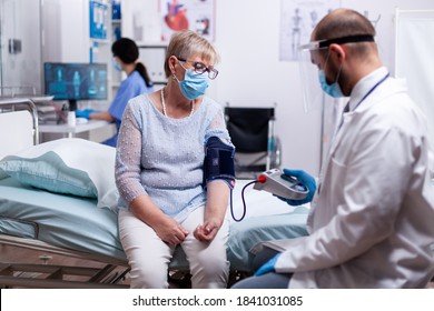 Doctor With Face Mask Against Coronavirus Taking Blood Pressure Of Senior Woman In Hospital Examination Room. Medical Control For Infections, Disease And Diagnosis.