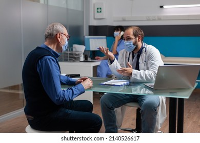 Doctor With Face Mask Consulting Senior Adult In Medical Office During Pandemic. Professional Medic Explaining Health Care Diagnosis And Recovery To Mature Patient At Checkup Appointment.