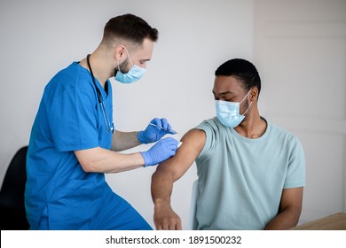 Doctor In Face Mask Giving Covid-19 Vaccine Injection To Black Male Patient During Vaccination Campaign At Clinic. Young African-American Man Getting Immunized Against Infectious Disease At Hospital