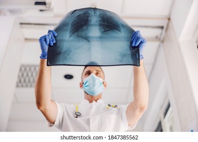 Doctor with face mask and rubber gloves standing in hospital and looking at x-ray of lungs during corona virus. - Powered by Shutterstock