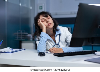 Doctor Falling Asleep At Medical Office After Hours, Working Late At Night. Exhausted Medic Looking At Computer Screen, Having Head In Hand Doing Overtime Work For Healthcare Job