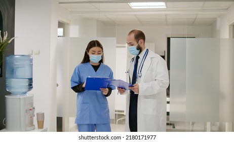 Doctor And Female Patient With Face Masks Talking In Waiting Room At Medical Facility, Doing Checkup Examination During Coronavirus Epidemic. Hospital Reception Covid 19 Prevention.