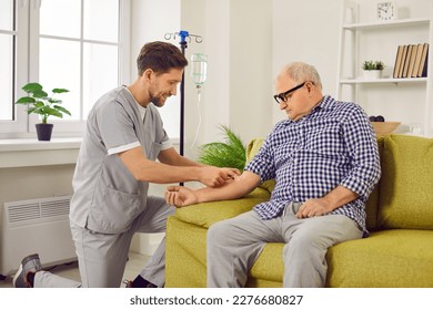 Doctor gives intravenous infusion to senior patient. Young nurse in scrubs uniform inserts IV line needle in vein of old man sitting on sofa at home. Medicine, vitamin therapy, medication concept - Shutterstock ID 2276680827