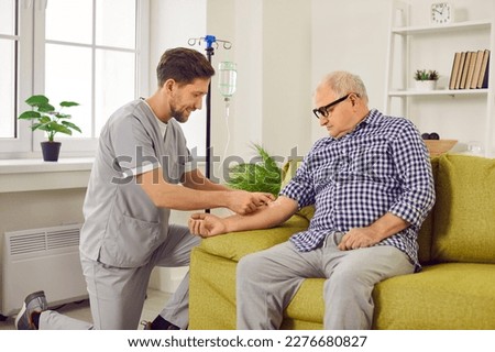 Doctor gives intravenous infusion to senior patient. Young nurse in scrubs uniform inserts IV line needle in vein of old man sitting on sofa at home. Medicine, vitamin therapy, medication concept