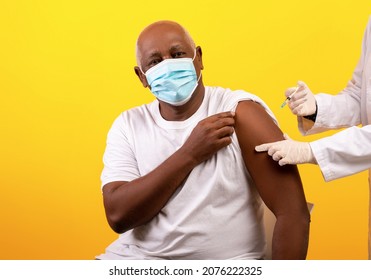 Doctor Giving Coronavirus Vaccine Shot To Senior African American Male Patient On Orange Studio Background. Elderly Black Man In Face Mask Being Immunized Against Covid-19