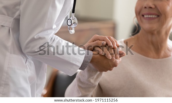 Doctor giving hope. Close up shot of young female
physician leaning forward to smiling elderly lady patient holding
her hand in palms. Woman caretaker in white coat supporting
encouraging old person