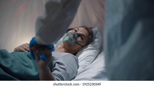 Doctor In Gloves And Ppe Suit Holding Hands Of Sick Covid-19 Aged Lady Lying In Bed At Quarantine Room. Medical Staff Supporting Ill Senior Woman Patient With Oxygen Mask In Hospital Ward