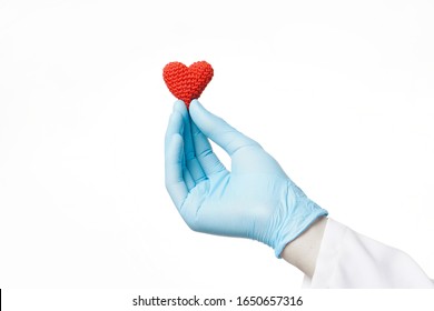 Doctor Hand In Blue Glove Holding A Crochet Red Heart On A White Background. Cardio Care And Love, Help With Heart Disease Medical Concept