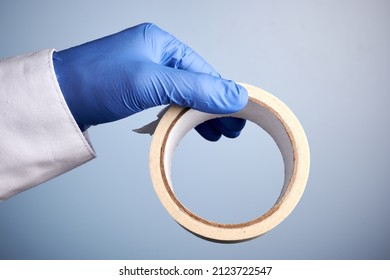 Doctor hand holds masking tape. Photo with blue background. Hand in blue nitrile glove. Adhesive paper scotch. Sticky tape roll. Gloved palm offers glue paper tape. Nurse hand with masking tape roll.