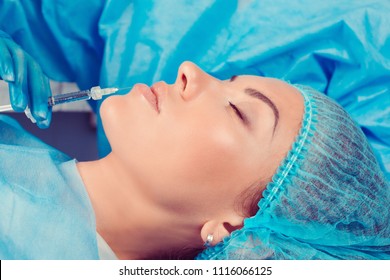 Doctor Hand Makes A Cosmetic Injection In The Lower Lip In A Clinic Of A Patient In Profile With Eyes Closed In A Surgery Room; O.R Background. Beautify Filler For Face Lips Concept