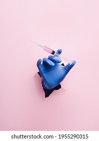 Doctor hand with surgical gloves breaking through the wall and holding syringe on pink background. Creative mass vaccination concept. Covid-19, influenza or flu global pandemic immunization.