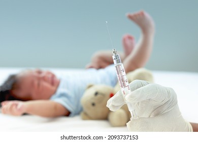 Doctor hand wearing medical glove and holding syringe with blurred background of little newborn infant baby lying down and crying, vaccinating baby, giving child vaccine injection concept.