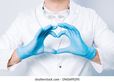 Doctor Hands In Gloves In The Heart Shape Against The Background Of His Body And Medical Gown. Closeup. Male Hand In Blue Medical Gloves Show Heart Sign