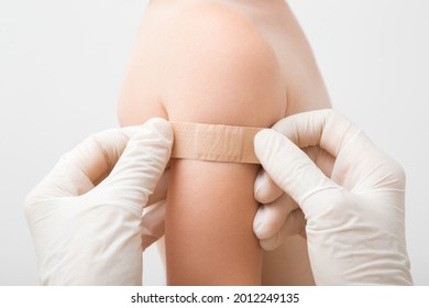 Doctor Hands In Rubber Protective Gloves Putting Adhesive Bandage On Baby Shoulder After Scratch On Skin Or Syringe Injection Of Vaccine. Patient Receiving First Aid. Closeup. Children Vaccination.