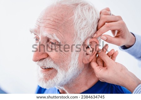 Doctor helps mature male patient to use hearing aid. Close up photo of a senior man smiling while using hearing aid. Deafness treatment concept
