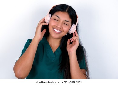 Doctor hispanic woman wearing surgeon uniform over white background smiles broadly feels very glad listens favourite music track via wireless headphones closes eyes.
