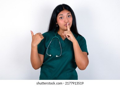 Doctor hispanic woman wearing surgeon uniform over white background asking to be quiet with finger on lips pointing with hand to the side. Silence and secret concept.