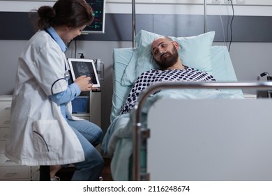 Doctor Holding Digital Tablet With Brain Mri Pointing At Results Indicating Tumor Explaining Treatment Options To Patient. Middle Aged Man Recieving Diagnosis While Connected To Oxygen Dispenser.