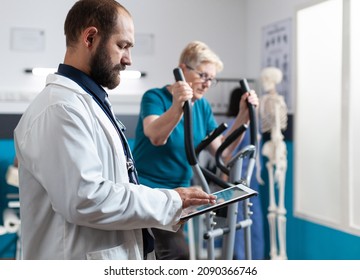 Doctor holding digital tablet for checkup with woman at recovery. Senior patient using stationary bicycle for physical exercise while talking to medical specialist about physiotherapy - Powered by Shutterstock