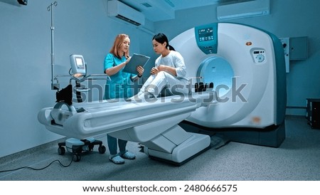 Doctor holding folder and making notes of patient symptoms. Patient lying at CT scanner. Modern equipped cabinet in private clinic. Doctor talking to patient before procedure. MRI examination.