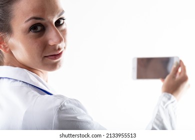 Doctor Holding Up Her Mobile Phone And Looking Back At The Camera Over Her Shoulder In A Cropped Portrait With Focus To Her Face