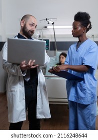 Doctor Holding Laptop Unhappy With Patient Lab Results Looking With Disapproval At Nurse Holding Clipboard In Hospital Ward. Physician Discussing Medical Treatment With Health Care Worker.