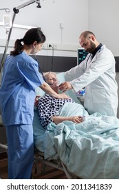 Doctor Holding Oxygen Mask For Senior Woman Patient Helping Her Breath, In Hospital Room With Respiratory Failure. Nurse Using Stethoscope Listening Sick Elderly Heart In Clinic. Iv Drip Bag Attached.