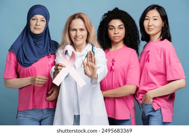 Doctor is holding a pink ribbon and making a stop sign with her hand. Surrounded by diverse women wearing pink to raise awareness for breast cancer. They are standing in front of a blue background - Powered by Shutterstock