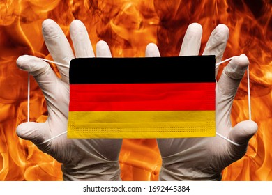 Doctor Holding In Protection Gloves Medical Face Mask, Respiratory Bandage With Germany National Country Flag Superimposed On Mask. Concept Travel Ban On Background Of Dangerous Red Flames Fire