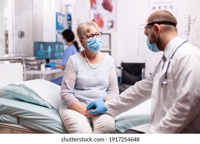 Doctor Holding Senior Woman Hand In Hospital Giving Compassion In Time Of Covid19. Medical Staff Wearing Surgical Face Mask Talking With Elderly Patient During Coronavirus Pandemic.