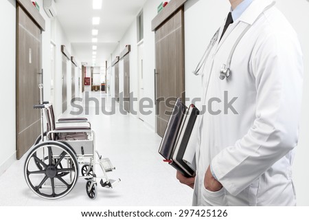  Doctor holding treatise book at empty wheelchair on corridor of hospital 