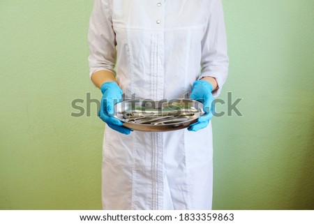 Doctor holds a kidney shaped tray with manicure and pedicure equipment, such as tweezers and scissors. Medical worker is dressed in white gown and blue latex gloves. Green wall background. 